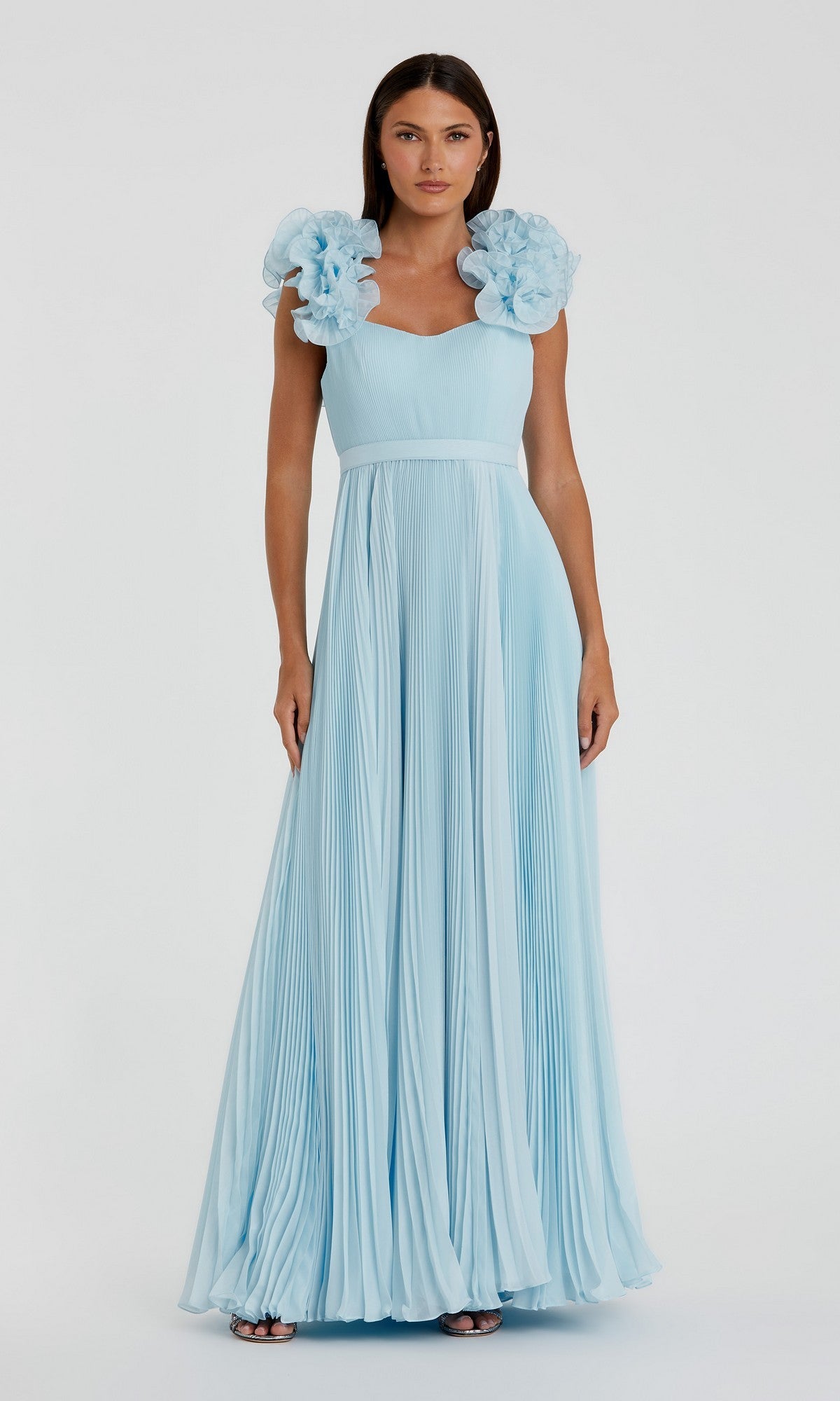 A woman stands facing the camera against a plain background, wearing the Mac Duggal 49979 long formal dress in powder blue, featuring floor-length pleated organza and prominent ruffled shoulder accents.
